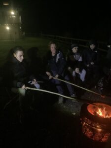 A group of students enjoy being around the camp fire by chatting and toasting marshmallows.