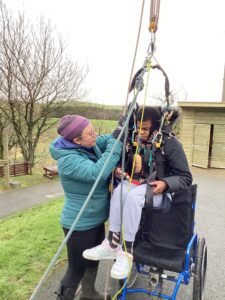 Safety first! A student is being lifted from a wheelchair in a harness as they prepare to go on the zip line.