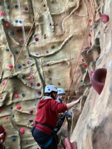 A student and member of staff are working hard to climb the climbing wall. The student is smiling and thoroughly enjoying the experience.